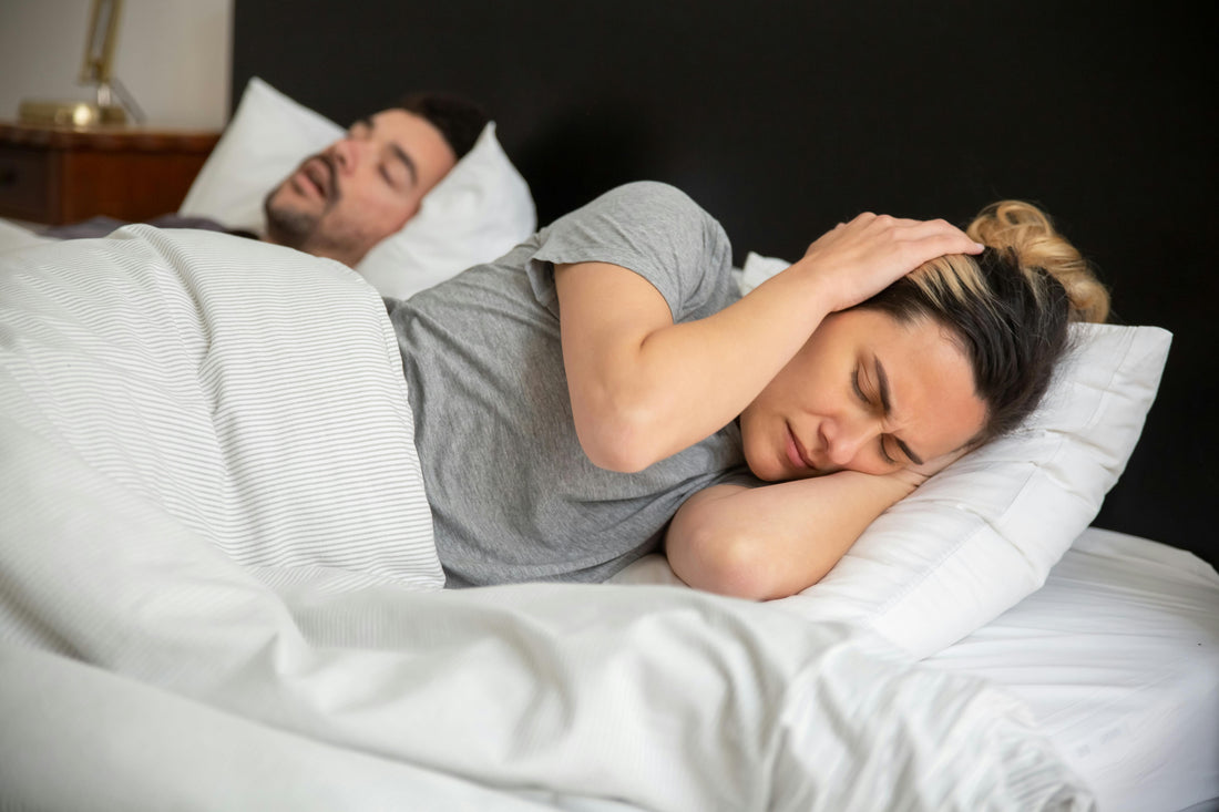 Woman covering her ear while her partner snores, illustrating disrupted sleep and the need for night-time noise reduction